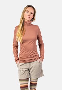 Bronzefarbener langer Pullover mit Rollkragen aus weichem Stoff, kombiniert mit beigen Cordshorts und mehrfarbigen gestreiften Leggings.