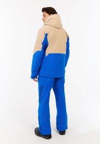 Mėlyna ir smėlio spalvos neperšlampama slidinėjimo striukė su kapišonu, su geometriniu raštu ir užtrauktukais kišenėse; derinama su derančiomis mėlynomis slidinėjimo kelnėmis.