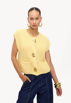 Gehaakt geel vest met korte mouwen, drie gouden knopen en twee voordezakken. Draagbaar over blauwe spijkerbroeken.