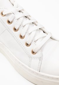 Baskets en cuir blanc avec des lacets blancs enfilés dans des œillets en métal doré et une semelle en caoutchouc blanche.