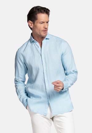 Man die een lichtblauw overhemd met lange mouwen en witte broek draagt, met één hand in zijn zak en naar de zijkant kijkt.