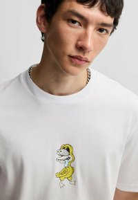 Hvid bomulds T-shirt med et tegneserie-dinosaur grafik i gul og sort, med teksten "HUGO" nedenfor karakteren på forsiden.