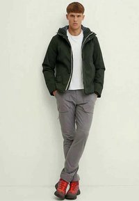 Young man wearing a black hooded jacket, white t-shirt, gray cargo pants, and red hiking boots standing against a plain wall with hands in pockets.