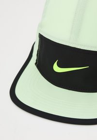 Lime zelena kapa s crnim remenom, s reflektirajućim žutim Nike logotipom. Izrađena od mekane tkanine s zakrivljenim vizorom i detaljima šivanja.