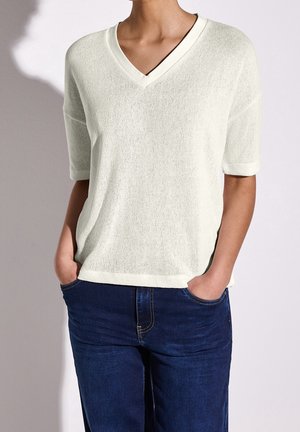 Hellbeiger kurzärmeliger Pullover mit V-Ausschnitt und strukturiertem Strickmuster. Kombiniert mit dunkelblauen Jeans, die Hände in den Taschen.