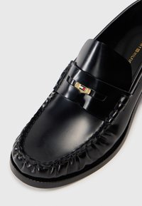Černé kožené mokasíny s kulatou špičkou, pletenými detaily a zlatými kovovými prvky s červeným akcentem. Hladký, lesklý povrch.