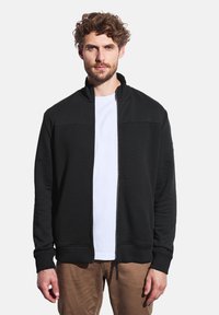 Sort lynlåsjakke med tekstureret kropspanel, høj krave og ribbede manchetter. Båret over en hvid t-shirt og kombineret med lysebrune bukser.