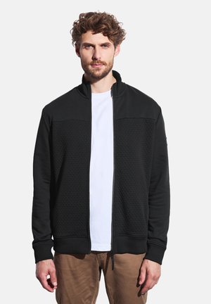 Zwarte zip-up jas met een textuur op het lichaam, hoge kraag en geribde manchetten. Gedragen over een wit t-shirt en gecombineerd met lichtbruine broek.