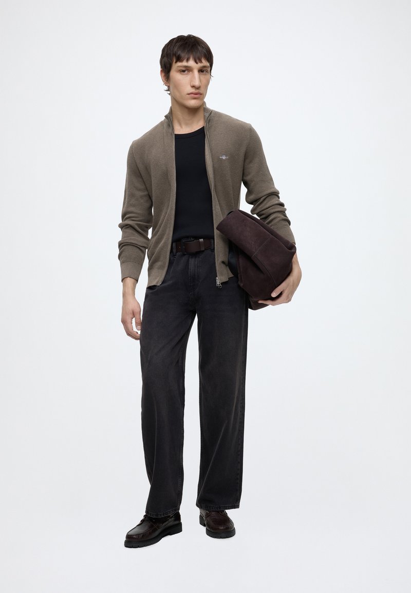 Joven con un suéter marrón con cremallera, camisa negra, vaqueros negros holgados y zapatos negros sosteniendo una bolsa marrón oscuro plegada sobre un fondo blanco liso.