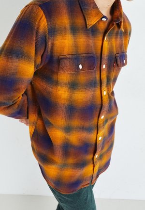 Mand iført orange og lilla ternet skjorte med knapper og forlommer, stående med hænderne bag ryggen mod en hvid baggrund.