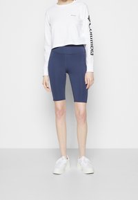 Bílá cropped košile s dlouhým rukávem a černým logem na rukávu, v kombinaci s námořnickými cyklistickými šortkami. Model nosí bílé tenisky. Hladká tkanina, přiléhavý design.