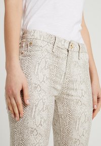 Jeans de estampado de serpiente en color beige con una textura suave, que cuentan con un cierre de botón, pasadores para cinturón y bolsillos traseros. Mano descansando en la cadera.