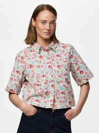 Jeune femme portant une chemise à manches courtes à fleurs rouges, roses et bleues, boutonnée, associée à un jean en denim foncé, mains dans les poches.