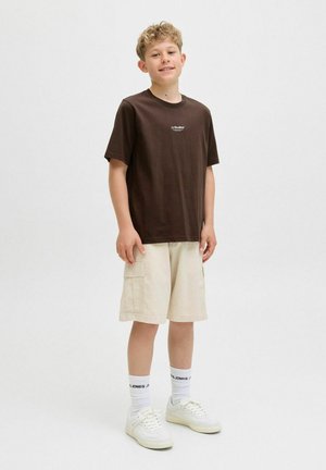 T-shirt in cotone marrone con un piccolo logo bianco, abbinato a pantaloni cargo beige e sneakers bianche con suola testurizzata e calzini con motivo.