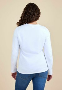Camisa de algodón blanca de manga larga con escote redondo, vista desde la espalda, combinada con vaqueros azules. Textura suave, ajuste regular.