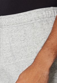 Šedé bavlnené tepláky s mäkkou textúrou, vybavené elastickým pásom a bočnou kapsou, na ktorých je vidieť časť ruky, ktorá spočíva na látke.
