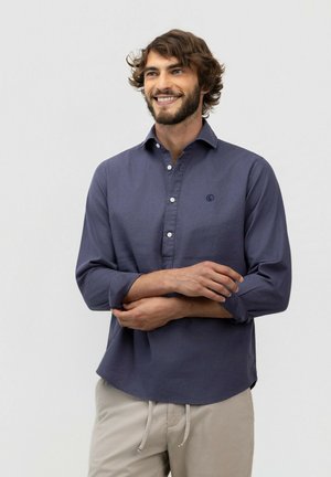 Hombre con barba que lleva una camisa azul oscuro abotonada y pantalones beige, sonriendo y sosteniendo un brazo con el otro frente a un fondo liso.