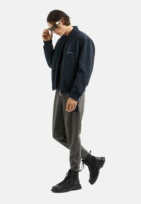 Granatowa kurtka bombowiec, szare dresowe spodnie, czarne wysokie buty. Kurtka posiada kieszeń z przodu, spodnie mają boczną kieszeń na zamek, elegancki design.