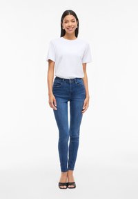 Camiseta blanca de algodón combinada con jeans ajustados azules. Los jeans tienen un diseño sencillo y bolsillos estándar. El calzado son tacones abiertos negros.