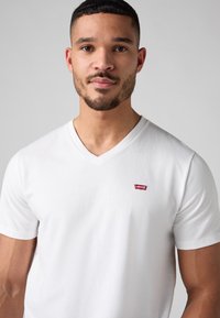 Valkoinen puuvillainen V-painikkeinen t-paita, jossa on pieni punainen Levi's-logo vasemmassa rinnassa, lyhyet hihat ja sileä pinta.