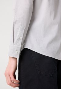 Camisa de botones a rayas con cuello, que presenta rayas verticales blancas y marinas; primer plano de la manga y una mano sosteniendo la tela.
