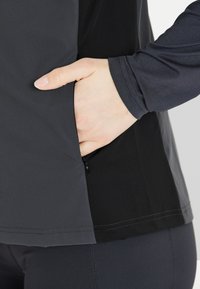 Langärmliges Oberteil aus schwarzem und dunkelgrauem Stoff, mit einer Reißverschlusstasche an der Seite und glatter Textur, außerdem mit gestepptem Saum und Nahtdetails.