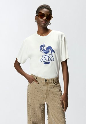 Žena koja nosi velike sunčane naočale u boji kornjačevine, bijelu majicu s plavom grafikom sirene i tekstom "Pinko Colada", bež hlače ukrašene zakovicama.