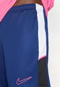 Ναυτικό μπλε αθλητικά σορτς με λευκή διχτυωτή επιφάνεια, ροζ λεπτομέρειες και ροζ λογότυπο Nike. Κατασκευασμένα από ελαφρύ, αναπνεύσιμο υλικό.