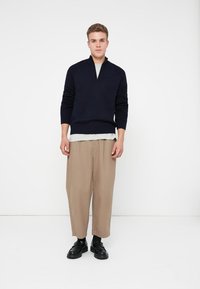Sweater de malha azul-marinho com meia abertura, sobre uma camisa cinza-claro, acompanhado de calças soltas bejes e sapatos pretos, de pé contra um fundo branco.