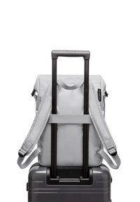 Sac à dos gris avec des bretelles ajustables posé sur une valise noire, dotée de deux poignées de tirage noires et d'une surface texturée.