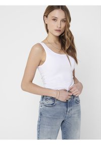 Débardeur blanc à encolure dégagée, associé à un jean clair déchiré. Le mannequin se tient avec les mains sur la taille, mettant en valeur une tenue décontractée.