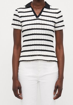 Persona vistiendo una camiseta de punto blanca de manga corta con rayas negras horizontales y un cuello negro, combinada con pantalones blancos.