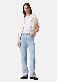Camiseta blanca de manga corta puesta debajo de un chal crema, combinada con jeans rectos de una tonalidad azul clara y calzado negro.