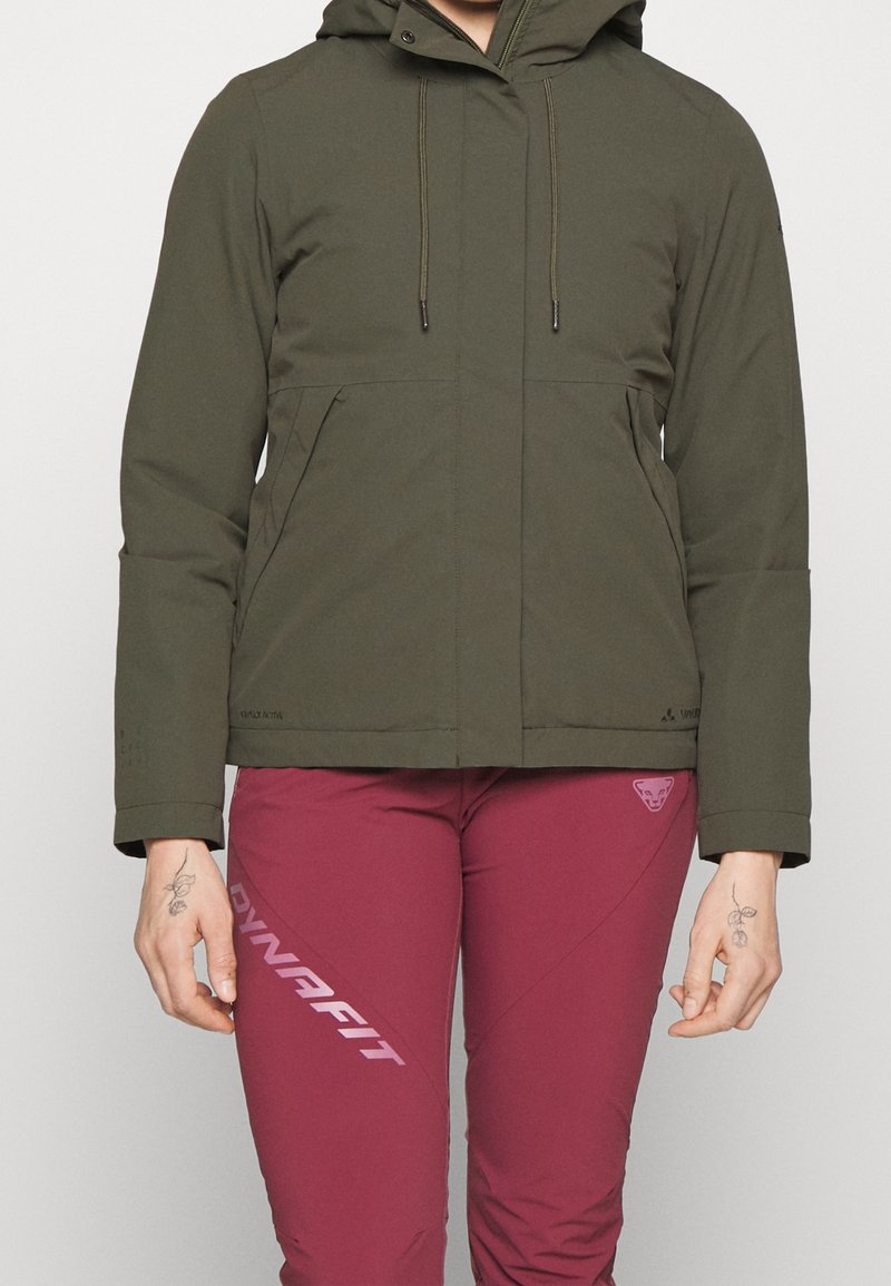 Personne portant une veste à capuche vert olive et un pantalon bordeaux de la marque Dynafit avec des tatouages de roses sur les deux mains.