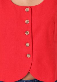 Red linen cropped top with a rounded neckline and five cream buttons along the front, featuring a smooth texture and curved hemline.