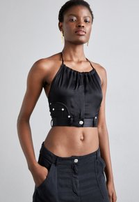 Čierny halter crop top zo saténovej tkaniny má zložený výstrih, bočné vrecká a strieborné cvoky. Hladká textúra a priliehajúci strih.