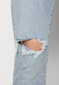 Lichtblauwe spijkerbroek met een grote, rafelige knie-scheur en kleinere slijtage-details, met een gladde textuur en een standaard pasvorm.