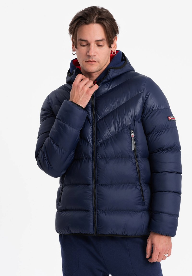 Marinblå puffjacka med huva, med chevron-quilting, dragkedjeförsedd ficka och kontrasterande röd innervadding. Snyggt, glänsande material.