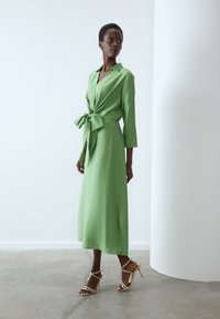 Groene, lange mouw wikkeljurk met een gestrikt taille-detail, gemaakt van textuurstof. Gestyled met gouden sandalen met hoge hakken.