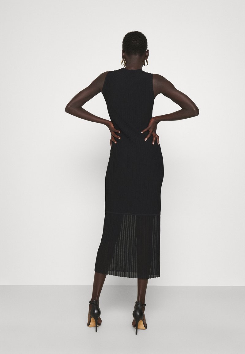 Femme portant une robe midi noire sans manches avec un ourlet plissé transparent et des sandales noires à talons hauts, debout les mains sur les hanches, dos face au spectateur.