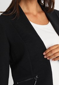 Czarna teksturowana marynarka z jedną kieszenią na zamek błyskawiczny, z kołnierzem szalowym i otwartym przodem, nałożona na biały top.
