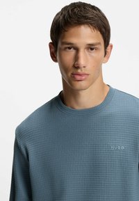 Hugo-merkkinen, teksturoitu svetari vaaleansinisenä, jossa on pyöreä kaula-aukko ja hillitty logopistely. Materiaali on pehmeän kudotun näköinen.