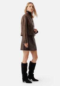 Giacca cropped in pelle marrone con bottoni neri, abbinata a una gonna coordinata. Stivali neri fino al ginocchio con dettagli frangi.