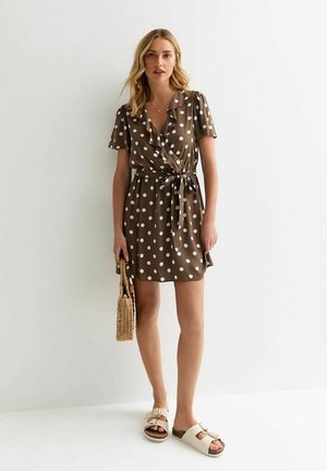 Vestido envolvente marrón con lunares, mangas cortas, cuello con volantes, cinturón en la cintura y un bolso de mimbre; combinado con sandalias de color claro.