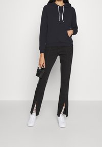 Marinblå huvtröja med vita dragsko, framficka, kombinerad med svarta jeans med sidofickor och vita sneakers, hållandes en liten svart väska.