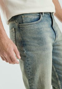 Man in een lichtblauwe vervaagde jeans en een beige shirt, met zijn linkerhand ontspannen naast zich.