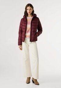Vrouw in een bordeauxrode pufferjack, met een roze-oranje gradient sweater, witte wijde broek en beige enkellaarsjes, staand voor een witte achtergrond.