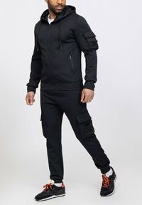 Haut à capuche zippé noir en tissu texturé, avec cordons de serrage ajustables et poches latérales ; pantalon cargo assorti avec poches ; chaussures de sport noires.