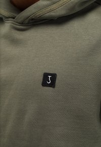 Dark green hoodie with a small black square patch featuring a white letter "J" sewn on the chest area.