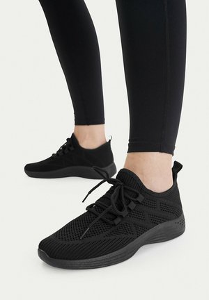 Osoba nosząca czarne, dziergane sportowe buty na sznurowadła i czarne legginsy, stojąca z jedną stopą lekko wysuniętą do przodu.